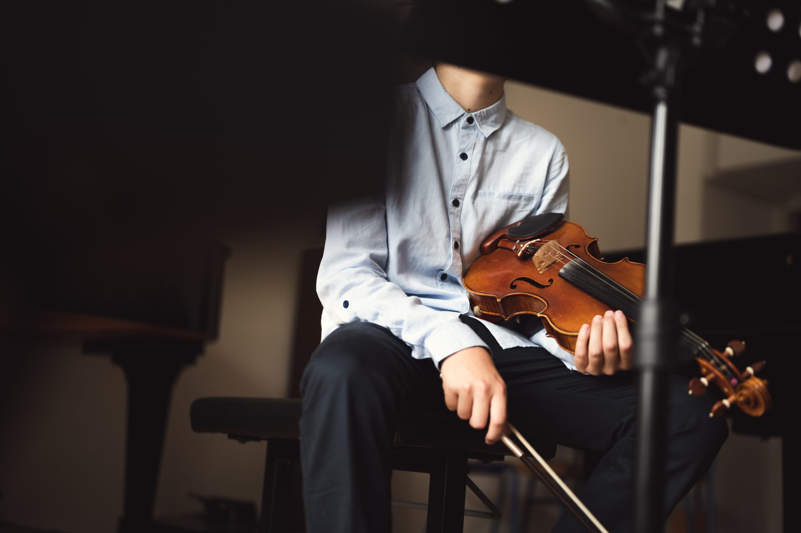 Unrecognizable teenage violinist sitting and holding instrument in his lap, resting between playing. Kid violin player indoors.